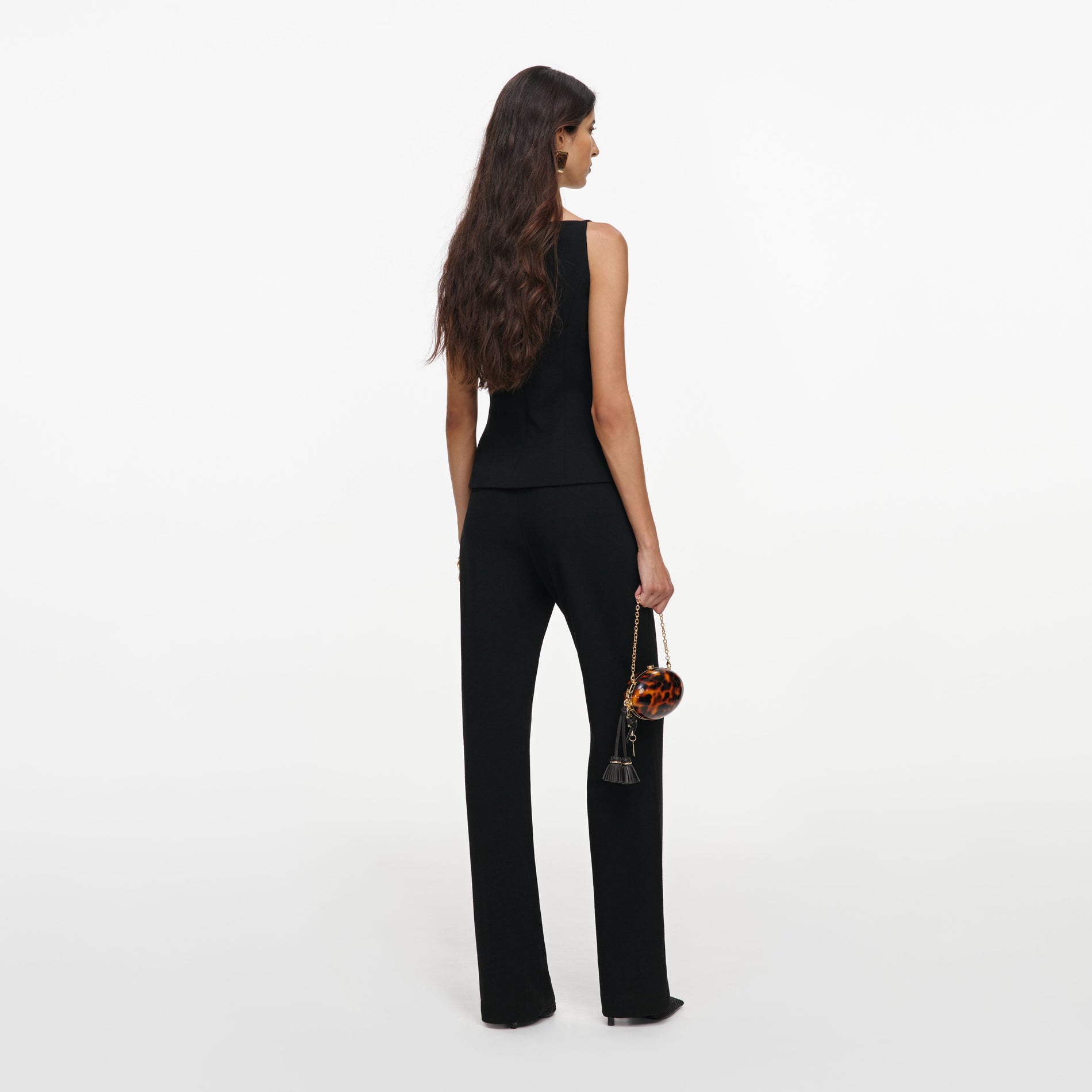 Woman in black sleeveless top and trousers, holding a tortoiseshell clutch bag with a tassel, viewed from behind