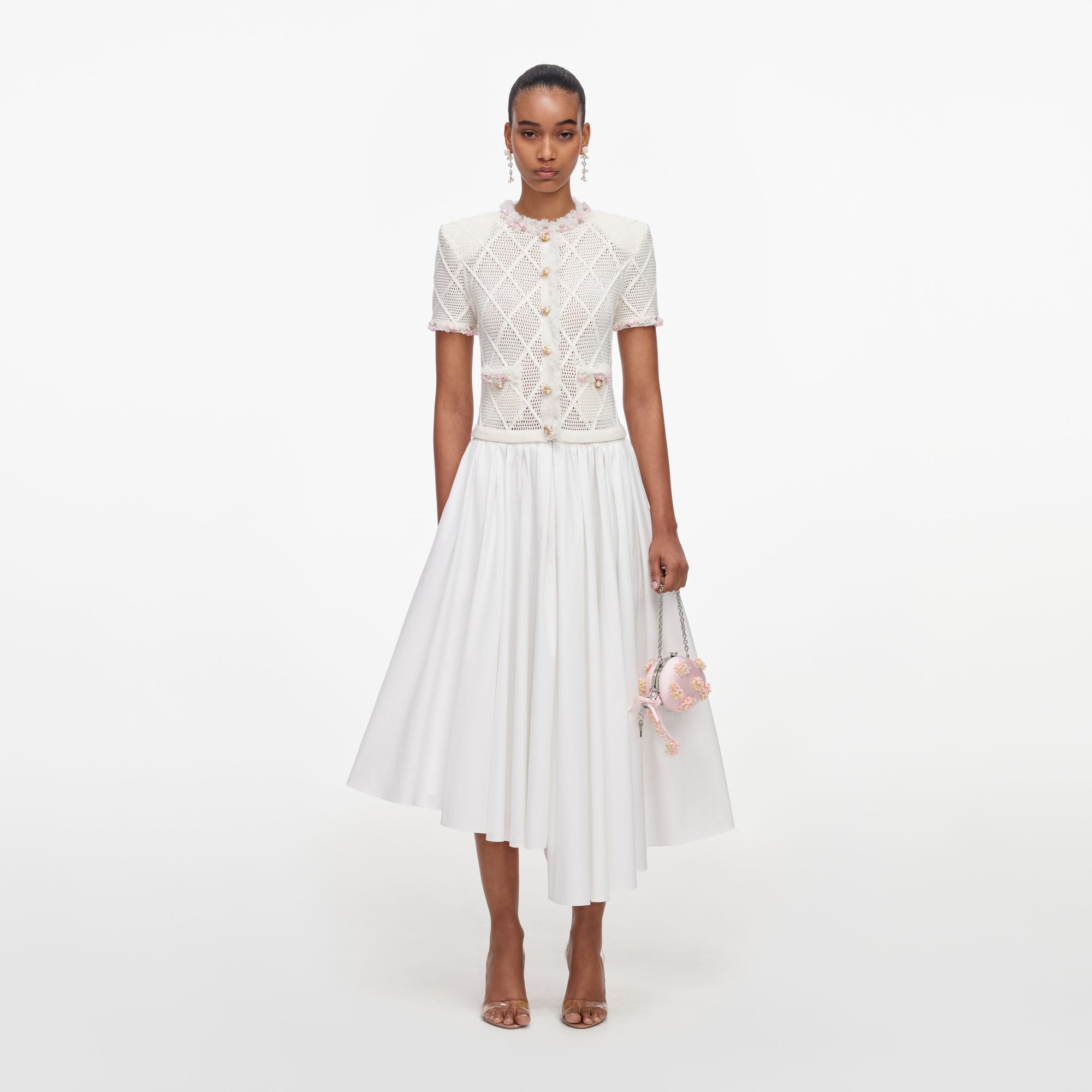 Woman in white short-sleeved top with pink trim, white midi skirt, clear heels, and pink floral handbag