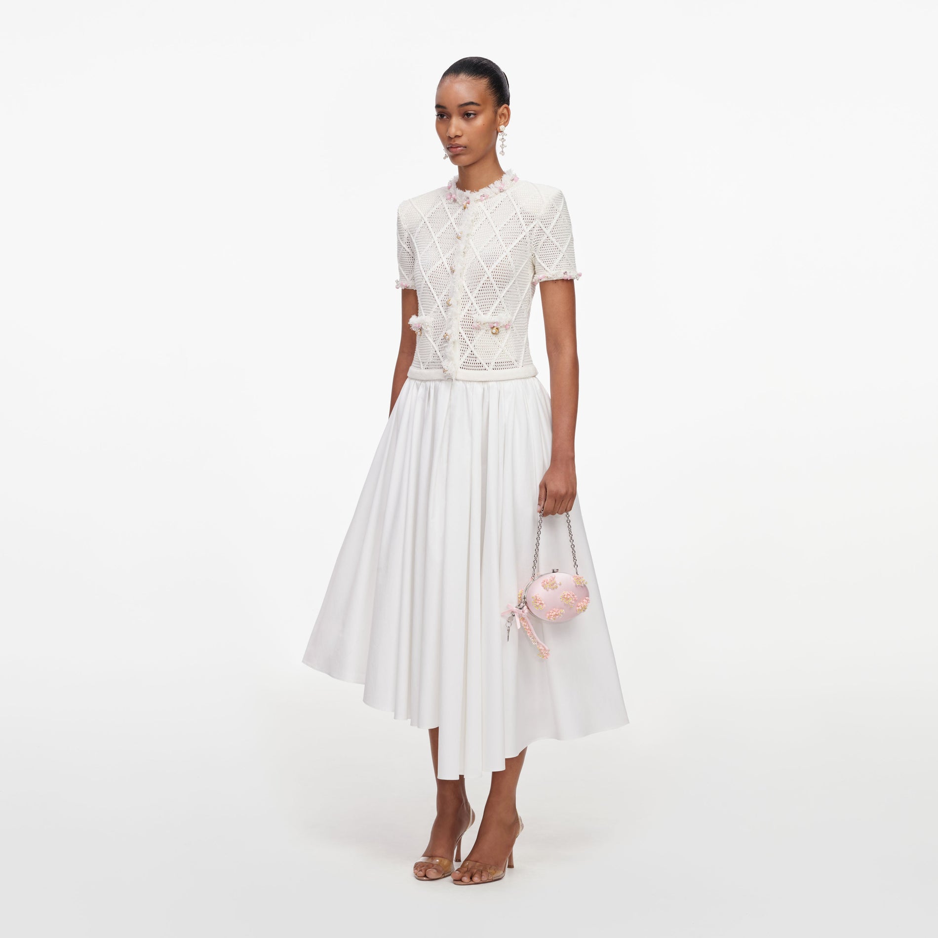 Model in a white short-sleeved crochet top with pink trim, a white midi skirt, and clear heels
