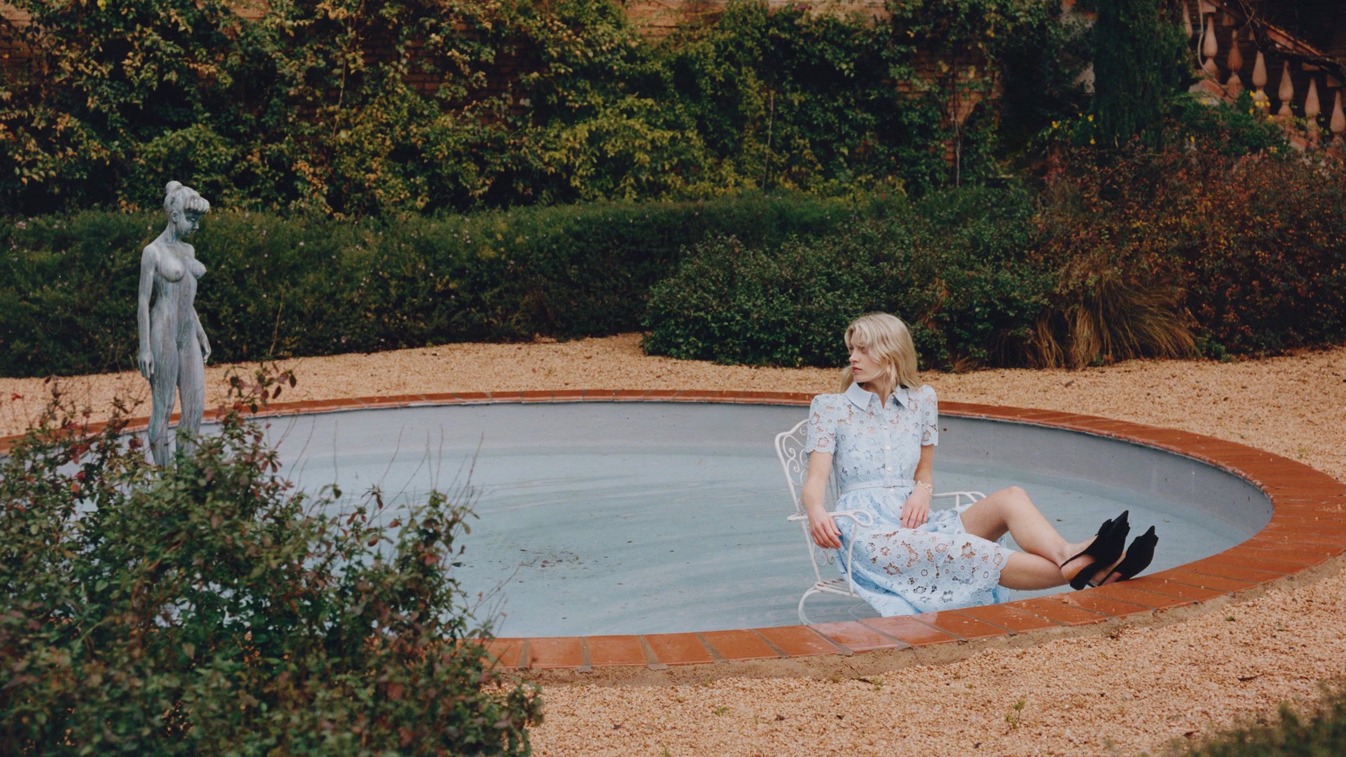 Woman in a blue lace dress sitting by a pool with a statue in the background.